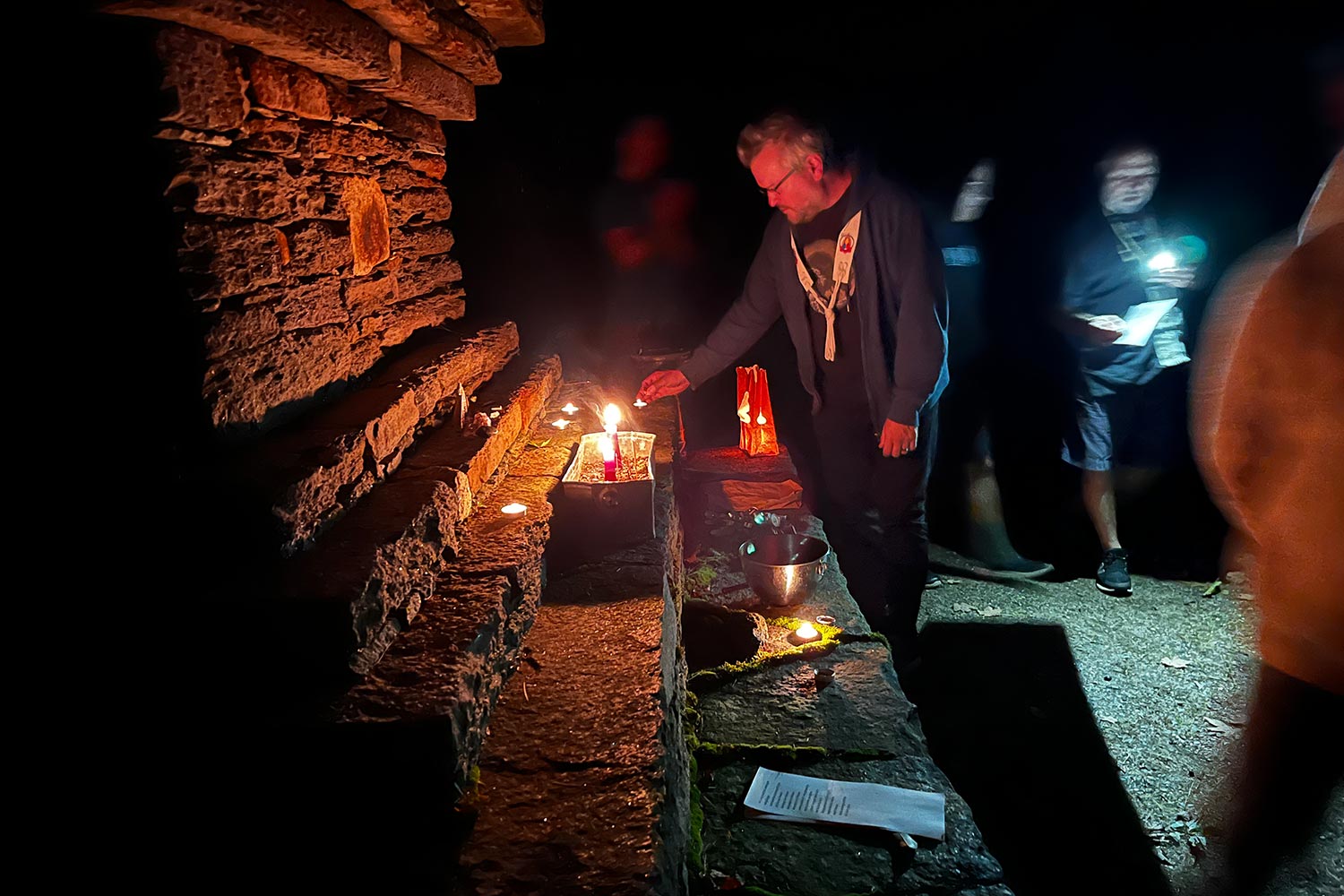 upcoming-retreats-retreat-1 A man in a ceremonial sash lights a candle at a rustic stone altar in the dark, surrounded by others holding lanterns and flashlights.