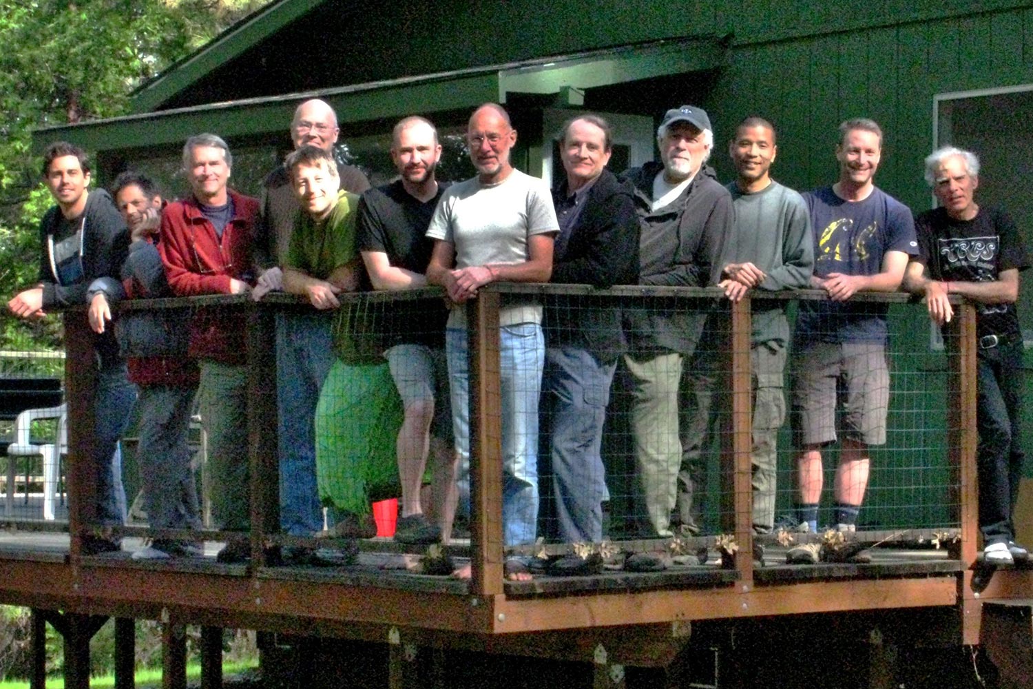 upcoming-retreats-retreat-2 A group of men standing on a wooden deck, leaning against a railing, smiling together with a cabin in the background.
